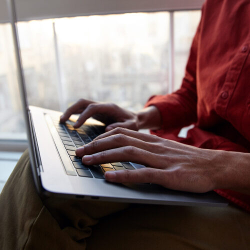 Cropped shot of young dark skinned guy dressed in red shirt and trousers keeping hands on keyboard while typing text, sitting near window on bright sunny day